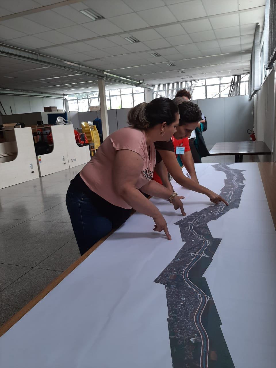 Imagem de Antonia, da SPObras, apresentando o trajeto do corredor, em uma pesa, com o dedo apontado para o percurso em questão e três pessoas em volta
